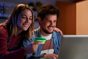Close up of a young couple doing online shopping from home at night. People using a laptop and a credit card to make a reservation.