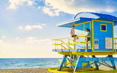 a couple on the beach in Miami Florida, a lifeguard hut in Miami beach, Asian women, and caucasian men on the beach during sunset. man and woman relaxing at a lifeguard hut looking at the ocean