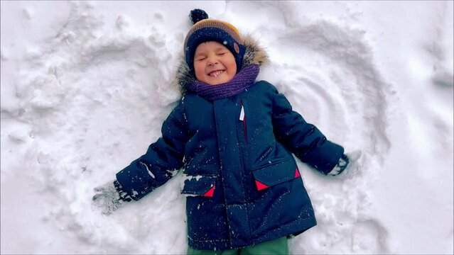 TOP view: Happy boy making snow angels. Flakes of snow fall on the child's face, he laughs and winces sweetly. Little boy playing in snowdrift and having fun with fresh snow. High quality 4k footage