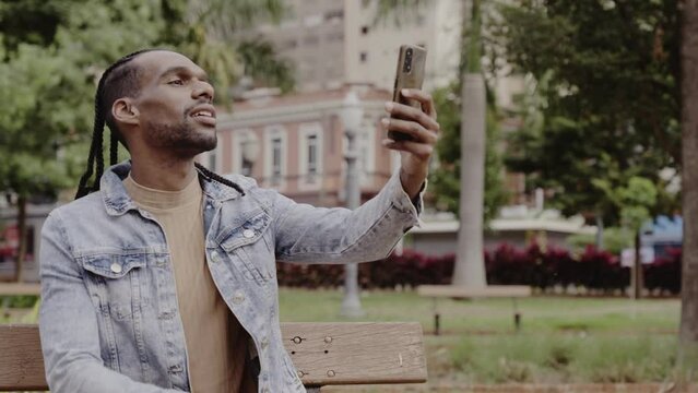 Jovem negro, sentado no banco da praca, sorrindo, tirando uma selfie com seu smartphone. Cinematico 4k.