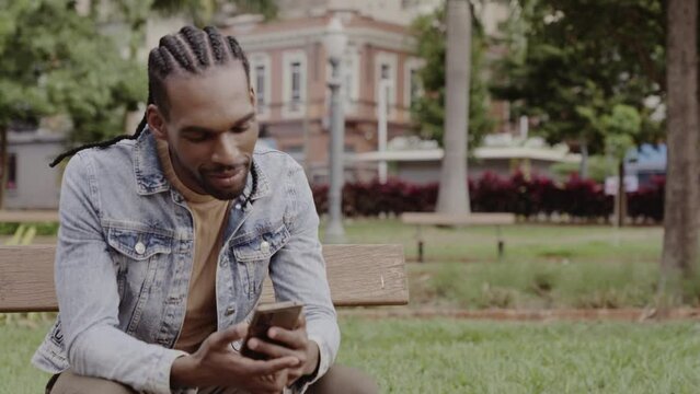 Jovem negro, sentado no banco da praca, sorrindo, usando seu smartphone. Cinematico 4k.