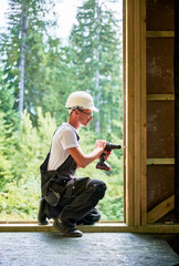 Carpenter constructing wooden framed house. Man worker in glasses working with screwdriver, wearing work overalls and helmet. Concept of modern eco-friendly construction.