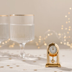 Time to New Year holiday celebration concept, gold vintage clock showing twelve o'clock, wineglasses with sparkling wine, blurred garland lights on background. Aesthetic neutral festive backdrop