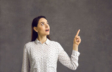 Wow, look here. Studio shot of surprised beautiful young woman with shocked face expression, big round eyes and open mouth looking and pointing finger up standing isolated on grey background