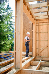 Carpenter constructing wooden framed house. Man worker cladding facade of house, using a screwdriver, wearing work overalls and helmet. Concept of modern eco-friendly construction.