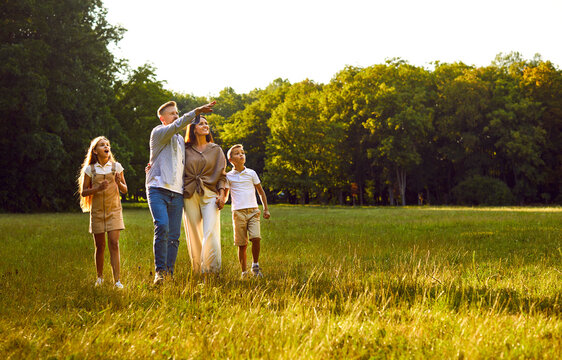 Happy family strolling in park in summer, walking on green grass lawn, enjoying beautiful landscapes, looking up, watching planes or birds flying in sky, relaxing and having good time together