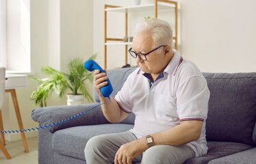 Senior man with dementia can't remember how to use the telephone. Confused puzzled old man in glasses with Alzheimer disease sitting on the sofa, looking at the phone receiver in his hand and thinking