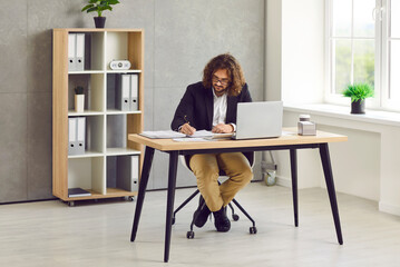 Portrait of a young attractive stylish business man with long hair working on a laptop at the...