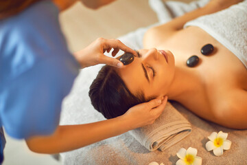 Close up portrait of a pretty young brunette woman with closed eyes lying relaxing in spa salon getting face and head massage therapy with hot stones. Wellness and beauty day concept. Top View.