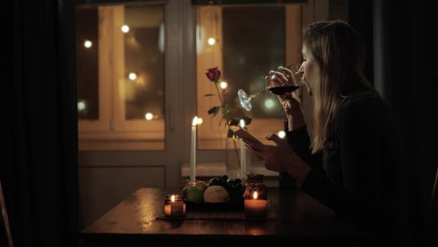 Working home, remote work. Attractive woman sitting alone at table in beautiful romantic setting by candlelight in evening with glass of wine and using phone, working