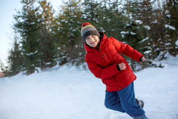 Fototapeta na wymiar Teenage boy in red puffed jacket and knitted hat has fun in the winter in the woods. Child pulls sled.