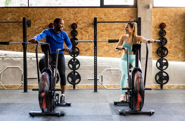 Front view of a couple of different ethnicities riding an air bike inside a gym. The young people...