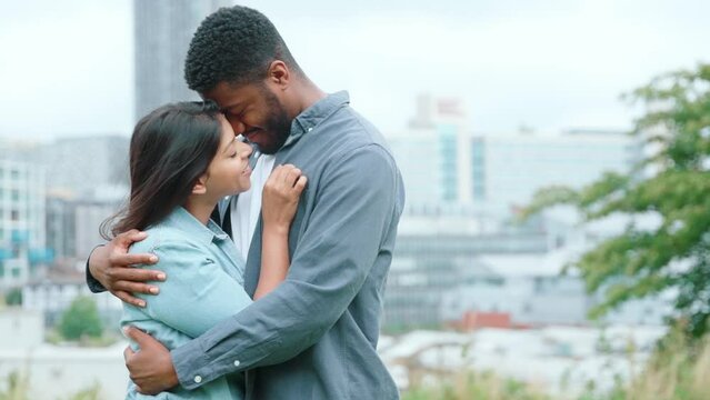 handsome african man and beautiful indian woman hugging each other in the park with a modern city in the background