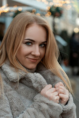 Portrait of a blond girl with beautiful eyes in a fur coat on a festive street. Christmas mood....