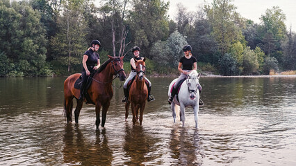 Three rider girls crossing the calm river water riding their beautiful horses on the cloudy summer...