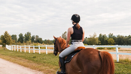 Female rider, horseback riding along the trail that leads between white wood fences and fields....