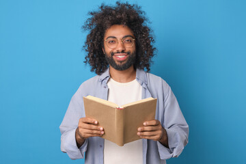 Young smart inquisitive Arabian man bookworm with glasses holding book urging to read more and gain new knowledge from literature stands in blue studio. Textbook, dictionary, encyclopedia