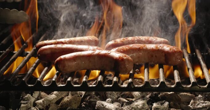 Flames seep through grill grate. Sausages are fried on hot grill. Cook lovingly turns sausages until they taste perfect. Fire coals give these delicious summer treats best flavor. Copy space, close-up