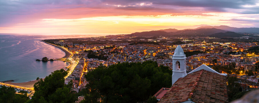 Panoramic photo from Sant Joan Hermitage, Blanes, with its white walls in the foreground and the illuminated city under a vibrant twilight sky.