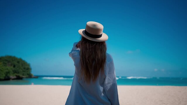 Romantic blonde woman tourist in straw hat walking to ocean sandy beach in sunny day, rear view. Female lady enjoying nature on vacations summer holidays. Travel, tourism, journey, rest relax concept.