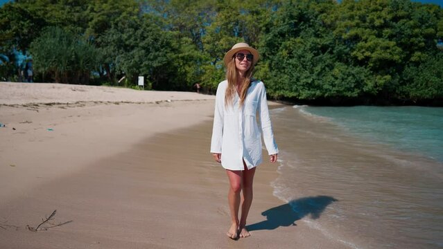 Woman in white airy tunic, straw hat walk on ocean sandy beach enjoy summer tropical vacations. Blonde female tourist rest by sea. tourism, travel.