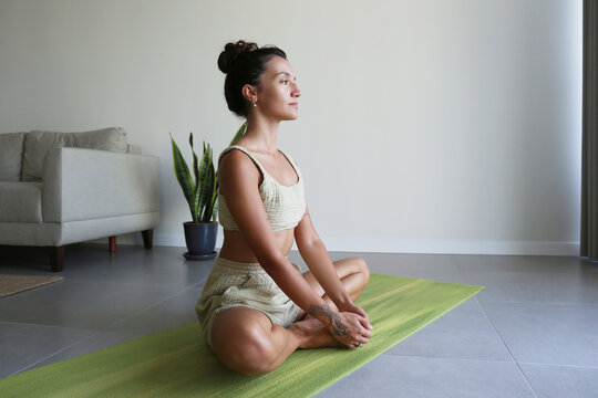Brunette young woman stretching in a butterfly pose, doing yoga at home. Fit middle eastern yogini performing an exercise for pelvic health at the living room. Close up, copy space, background.