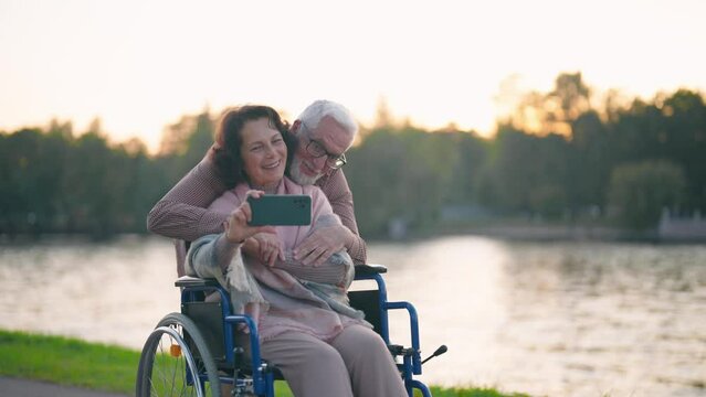 Wife in wheelchair makes couple selfie. Senior happy family spending time in city park near pond taking photos on smartphone. Man hugging woman showing love care. Living with physical disability.