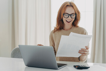 Joyful woman with glasses holding papers, celebrating mortgage closure near laptop