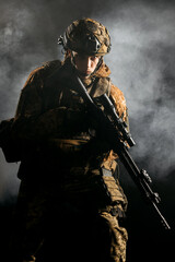 An army soldier in a military camouflage uniform, a helmet and a mask, holds a rifle and aims with a red dot sight, standing in an attack position, in the smoke on a black background.