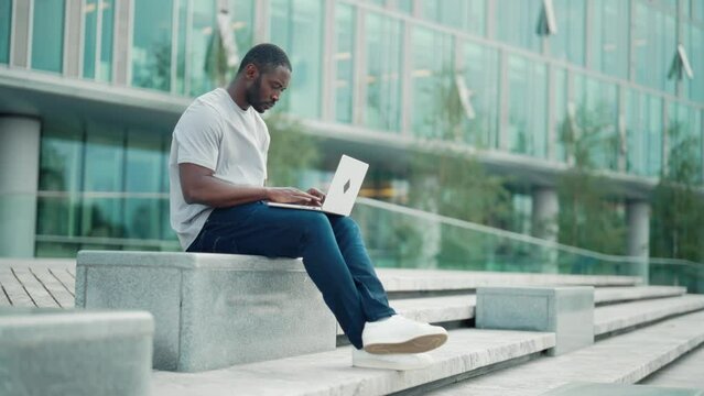 Serious focused man works on laptop sitting on street near modern office building outdoors. African american attentive guy freelancer thinking solving hard work problem online. Freelance work concept.