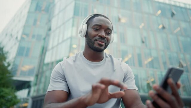African american man music lover in headphones enjoying listening music on smartphone sitting on city street outdoors. Positive smiling guy melomaniac rhythmically nodding head relaxing moving body.