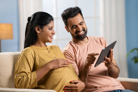 Happy Indian husband with pregnant wife discussing from digital tablet at home - concept of pregnancy preparations, togetherness and spending time