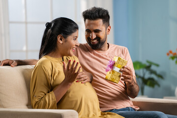 happy indian husband giving gift to pregnant wife by hugging at home - concept of romantic relationship, affection and maternity lifestyles.