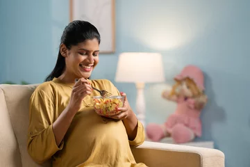 Schilderijen op glas Happy indian pregnant woman eating fruits salad while sitting on sofa at home - concept of healthy lifestyles, Maternal Pregnancy nutrition. © WESTOCK