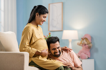 Happy indian husband listening baby sound of pregnant wifes belly at home - concept of Parental...