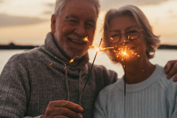 Couple of two old seniors holding sparklers lights at the evening at the beach together enjoying...