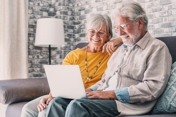 Pretty elderly 70s grey-haired couple resting on couch in living room hold on lap laptop watching...