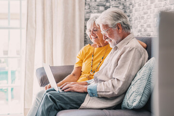 Pretty elderly 70s grey-haired couple resting on couch in living room hold on lap laptop watching movie smiling enjoy free time, older generation and modern wireless technology advanced users concept.