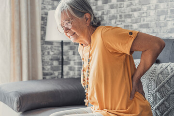 Unhappy mature woman touching back, feeling pain, sitting on couch in living room, unhealthy middle...
