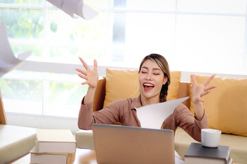 Happy beautiful Asian woman has fun by throwing papers into the air for celebrating business success or job done. Smiling young female student feels relaxed after finish studying of a test at home.