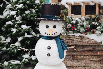 Snowman background. Christmas pine tree covered with snow. Smiling snowman with a scarf and carrot as a nose. Winter season background. Wooden cottage house in background.