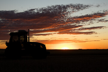 Tractor parked in a backlit field as the sun sets. Sky with clouds in orange tones. Copy space
