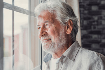 Happy thoughtful older 70s man looking out of window away with hope, thinking of good health, retirement, insurance benefits, dreaming of future. Elderly pensioner waiting meeting with family.