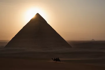 Foto op Canvas The sun dipping just behind the tip of the Great Pyramids of Giza as sunset settles on the ancient land of Egypt, creating a majestic and breathtaking view for tourist on camel rides passing thru. © Sweeann
