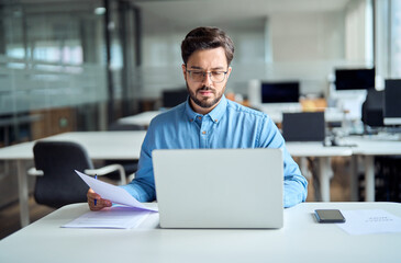 Busy latin young businessman checking document using computer in office. Young professional...
