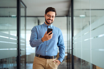 Smiling busy professional latin business man walking in office hallway holding mobile cellphone. Young happy businessman employee using smartphone looking at cell phone tech standing at work. - obrazy, fototapety, plakaty
