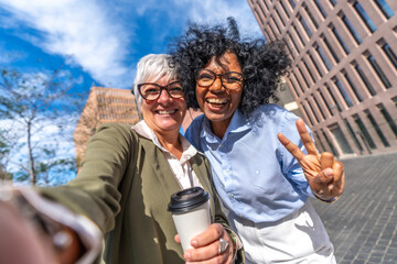 Smiling businesswomen taking selfie and gesturing victory with fingers outdoors