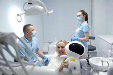 middle-aged woman in dental chair shows tongue and laughs she hides behind soft toy white astronaut in background doctor and nurse look at each other latest technology fun joy pleasure
