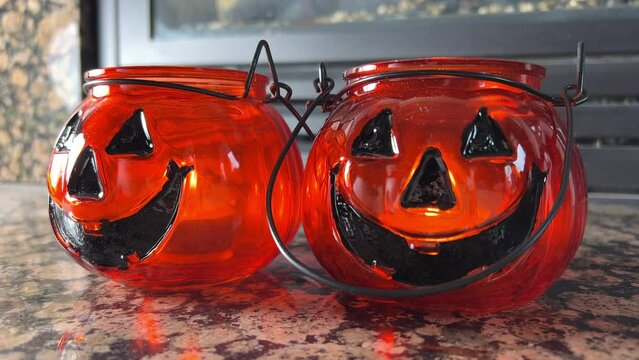 Two bright orange candlesticks in the form of pumpkins Jack-o-lantern Glass lantern with real candles Black eyes and black smile Transparent orange glass Standing in front of a fireplace