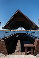 A wooden gazebo in the shape of a boat with a deck and a sail roof near the river